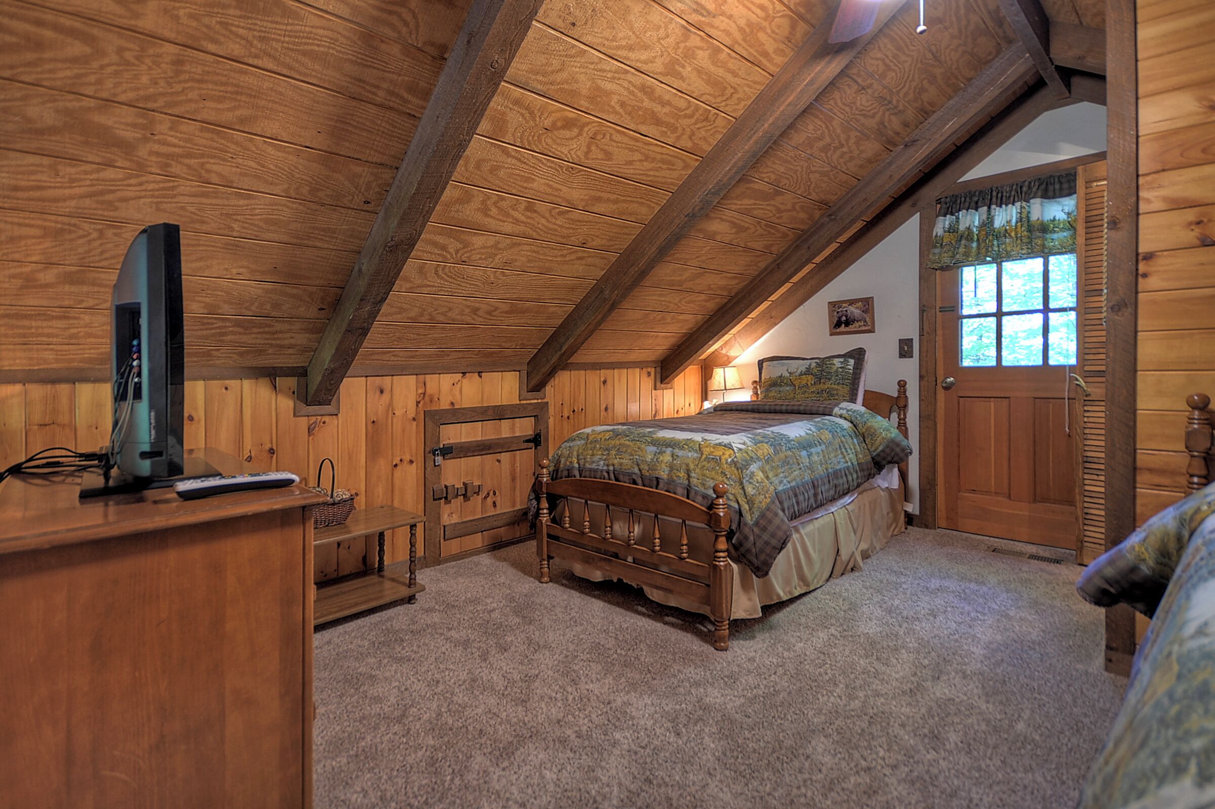 A bedroom with a wooden ceiling, a bed with a quilt, a wooden dresser, a television, and a door with a window.