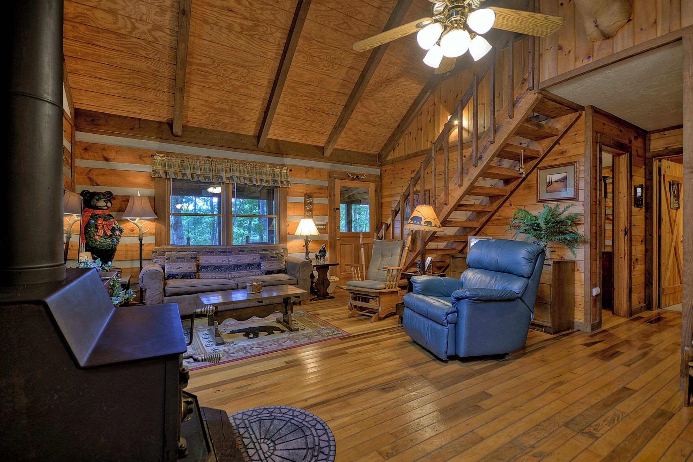 A wooden cabin interior with a fireplace, a sofa, a blue leather chair, and a staircase.