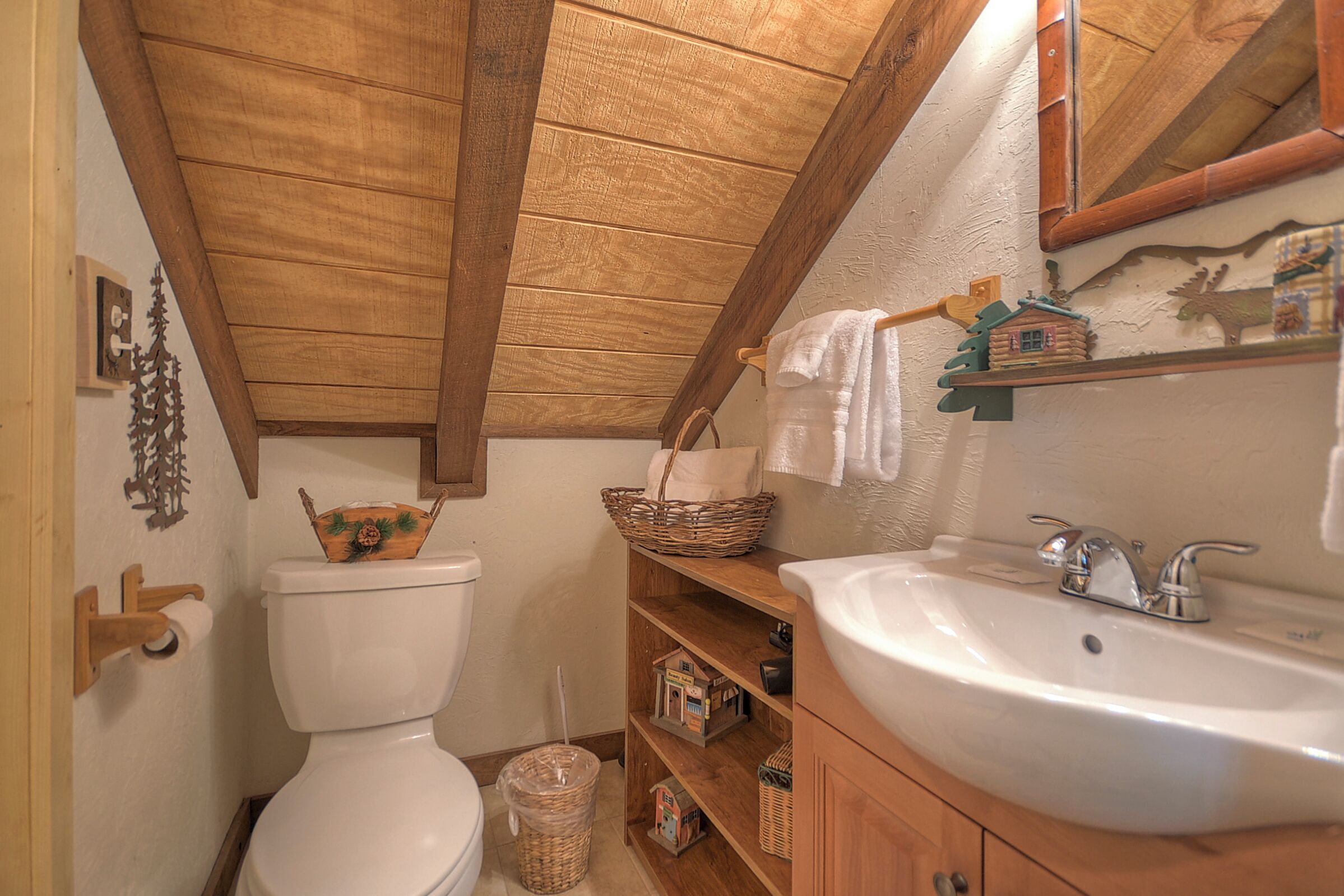 A bathroom with a wooden ceiling, a white toilet, a wooden shelf with a basket, a white sink, and a mirror.