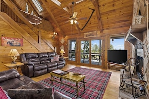 A log cabin interior with a leather couch, a plaid rug, a fireplace, and a television.