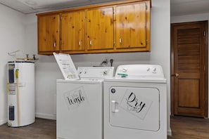 Une buanderie avec deux machines à laver et deux sèche-linge blancs, des armoires en bois au-dessus, et une porte en bois sur le côté droit.