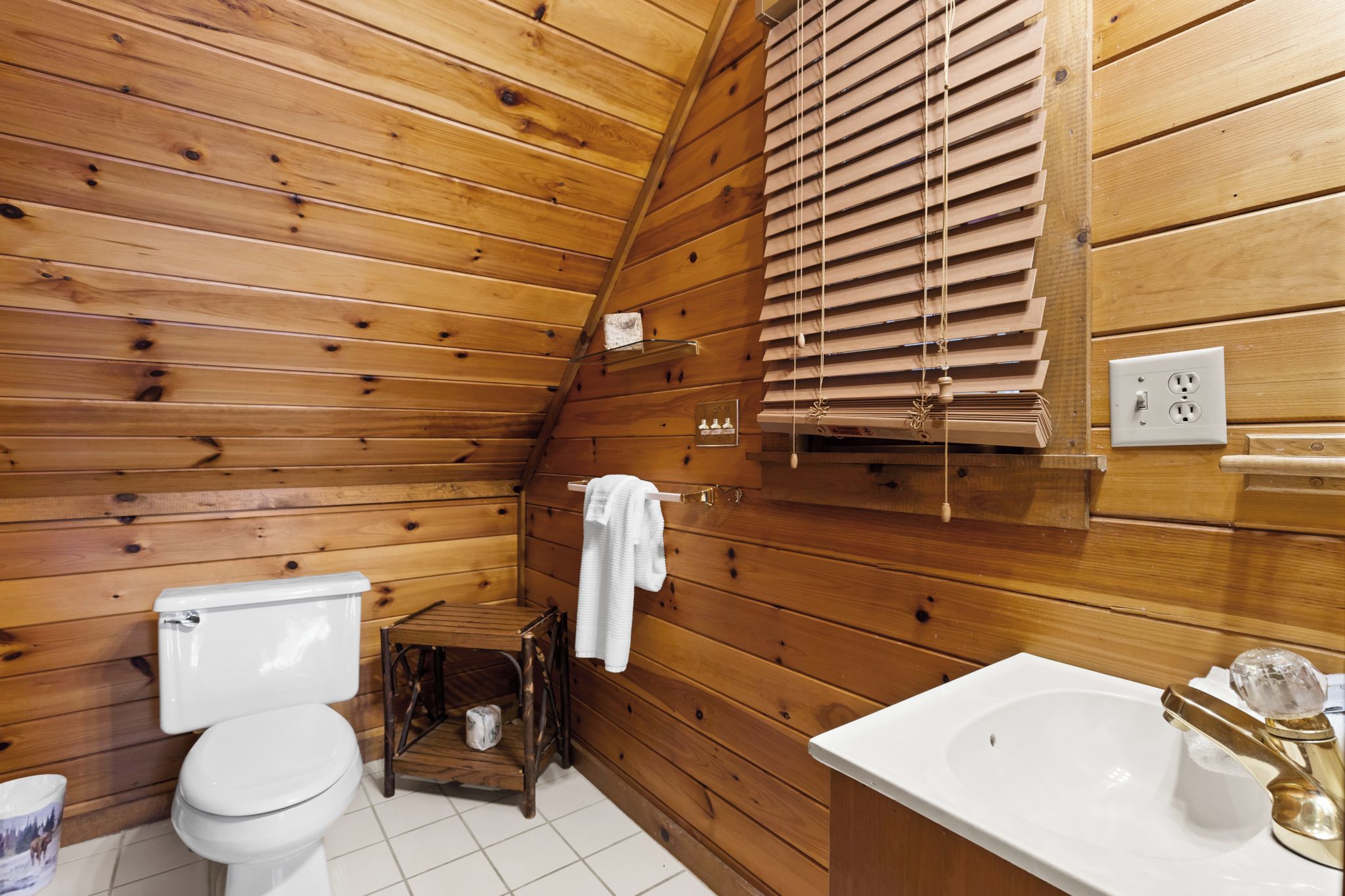 Une salle de bain aux murs lambrissés, équipée de toilettes, d’un lavabo et de stores aux fenêtres.