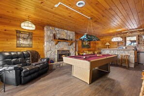 Un intérieur de chalet chaleureux avec une table de billard, un canapé en cuir, une cheminée en pierre et un plafond en bois.