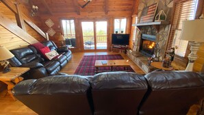 Un intérieur de chalet chaleureux avec une cheminée en pierre, des canapés en cuir, un téléviseur et un ventilateur de plafond.