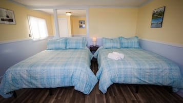 A room with two beds, wooden flooring, a nightstand with a lamp, and a framed picture on the wall.