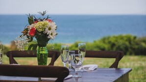 Une table dressée pour deux personnes, agrémentée d’un vase vert rempli de fleurs et de trois verres, avec vue sur une étendue d’eau.