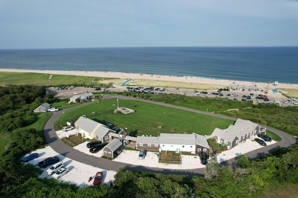 An aerial view of a coastal property with a large lawn, multiple buildings, and a parking area. - Nauset Beach Inn (Orleans)