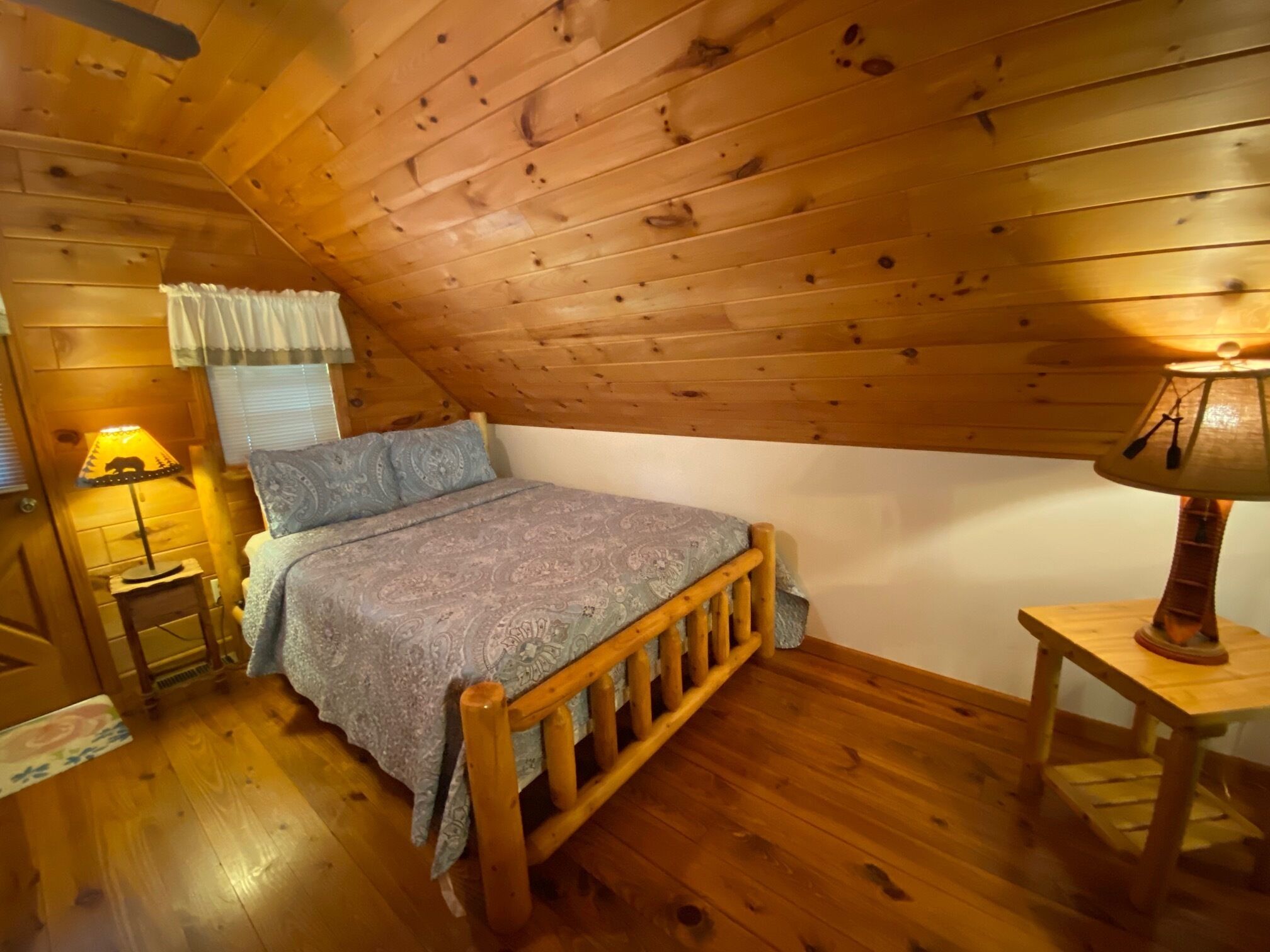 A cozy attic bedroom with a wooden sloped ceiling, a bed with a patterned bedspread, two bedside lamps, and a small wooden nightstand.