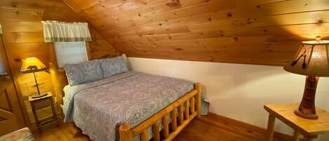 A cozy attic bedroom with a wooden sloped ceiling, a bed with a patterned bedspread, two bedside lamps, and a small wooden nightstand.