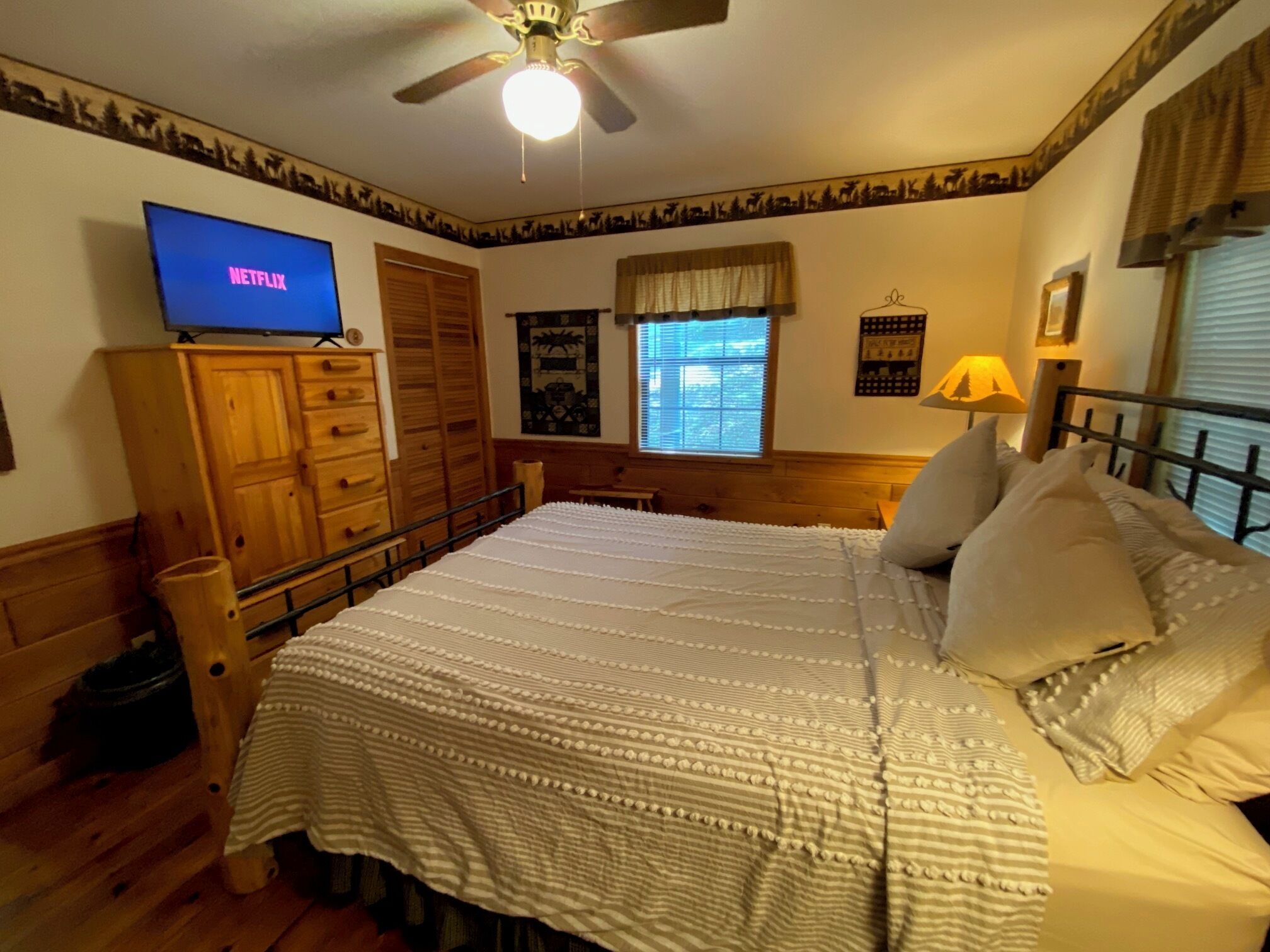 A bedroom with a bed, wooden wardrobe, a television displaying 'NETFLIX', a window with blinds, and a ceiling fan.