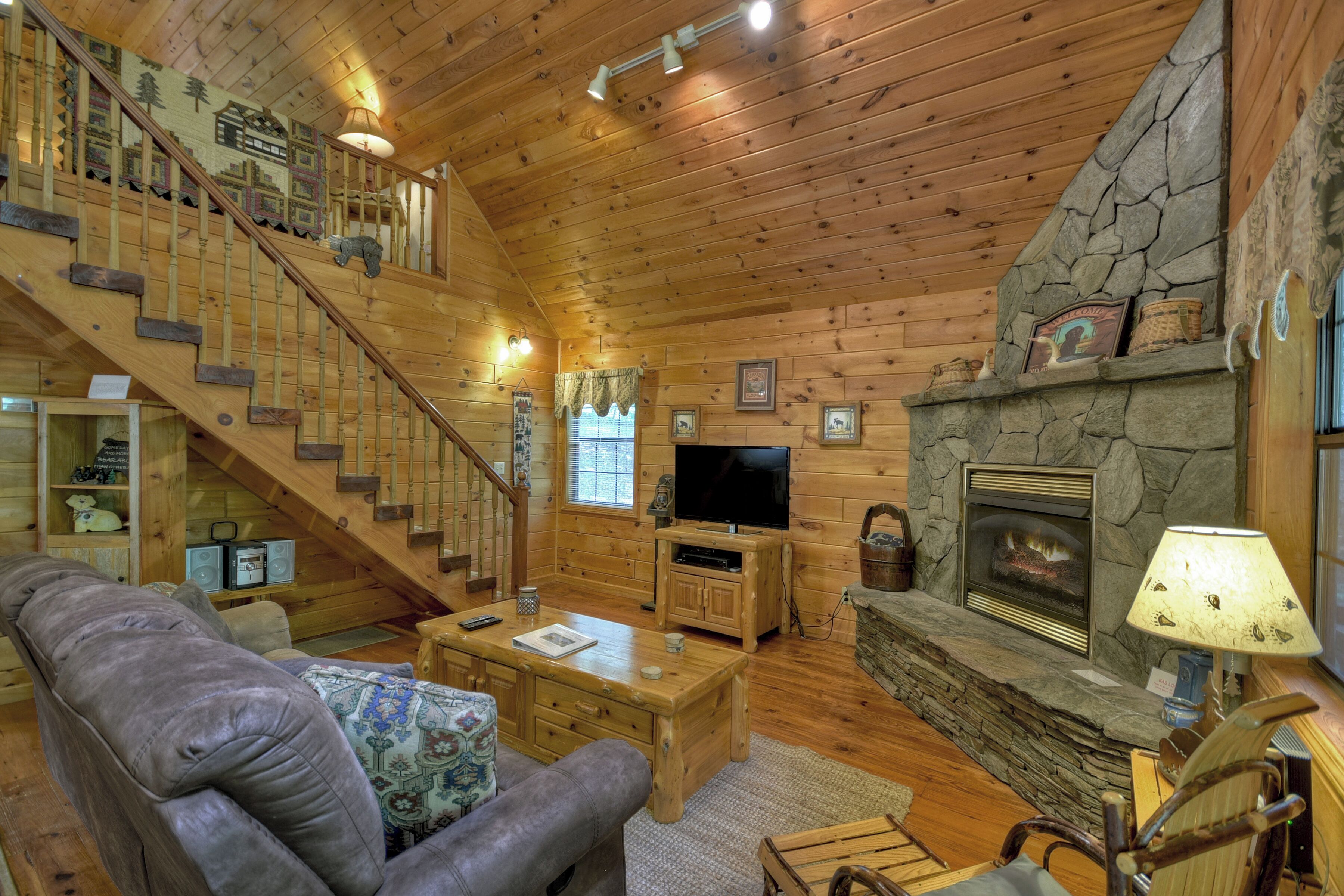 Un intérieur chaleureux de chalet en bois, avec un escalier en bois, une cheminée en pierre, un téléviseur et un coin salon confortable.
