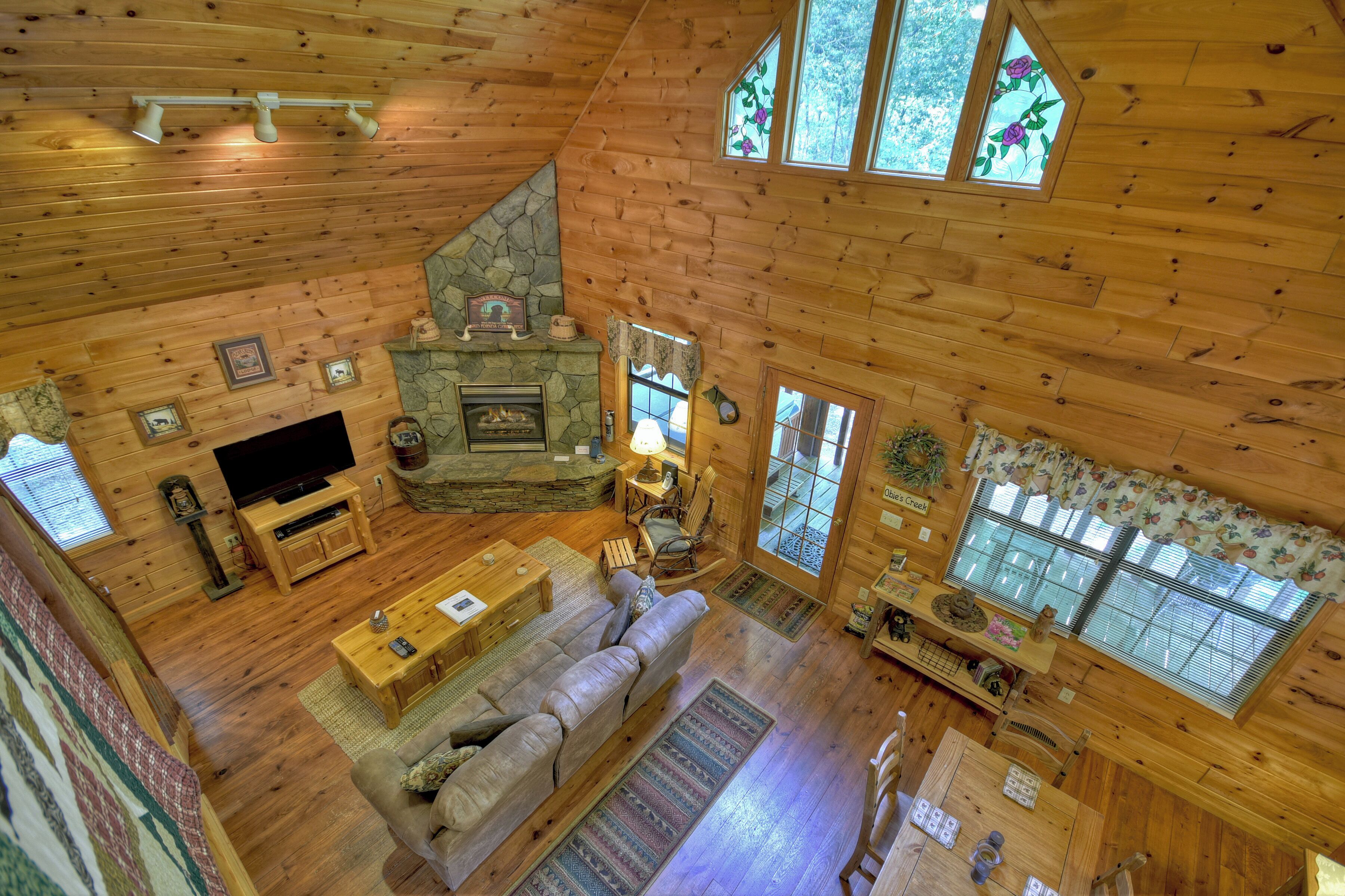 A log cabin interior with a stone fireplace, wooden furniture, and large windows.
