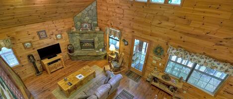 A log cabin interior with a stone fireplace, wooden furniture, and large windows.