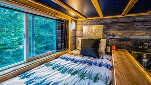 A bedroom with a large window, wooden ceiling beams, a bed with a forest-themed bedspread, a sign reading "NAP", and a nightstand with a lamp. - Fun Lofted Waterfront Home w/ Sauna and BBQ (Longview)