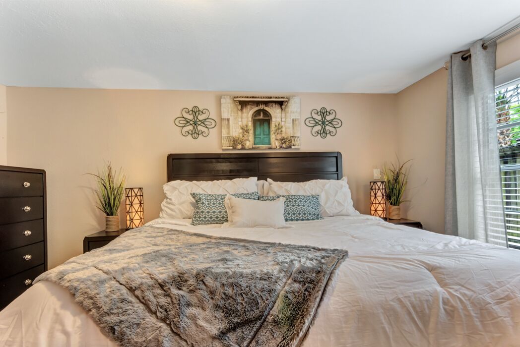 Um quarto com uma cama grande, duas mesas de cabeceira, cômoda, arte decorativa na parede e uma janela com cortinas.