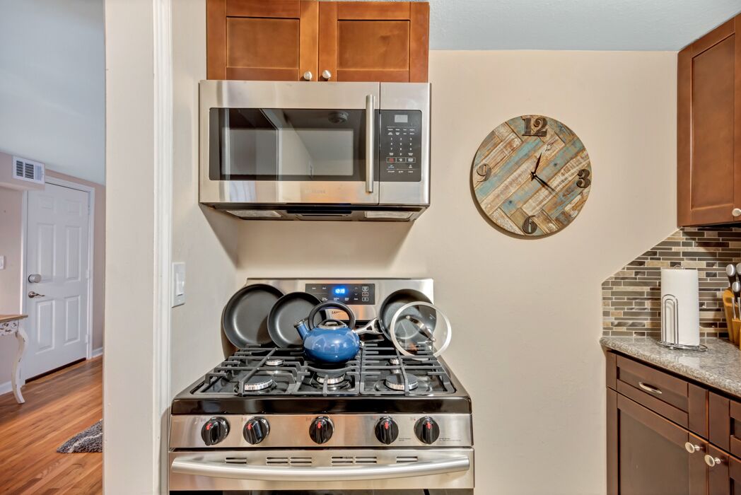 Une cuisine équipée d’un four à micro-ondes en acier inoxydable, d’une cuisinière avec des casseroles, d’une bouilloire bleue et d’une horloge rustique accrochée au mur.