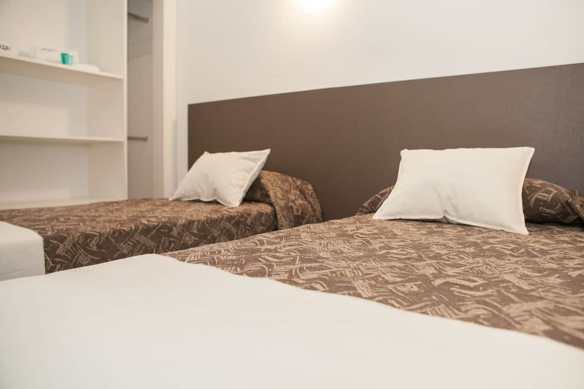 A hotel room with two beds, each with patterned bedding and white pillows. There is a white shelf unit with a few items on it.