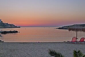 Uma praia com um pôr do sol, águas calmas e alguns barcos. Há cadeiras e um guarda-sol de palha na areia.