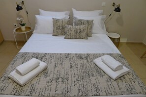 Uma cama bem arrumada com lençóis brancos, dois travesseiros decorativos com padrão caligráfico e duas toalhas enroladas sobre a cama.