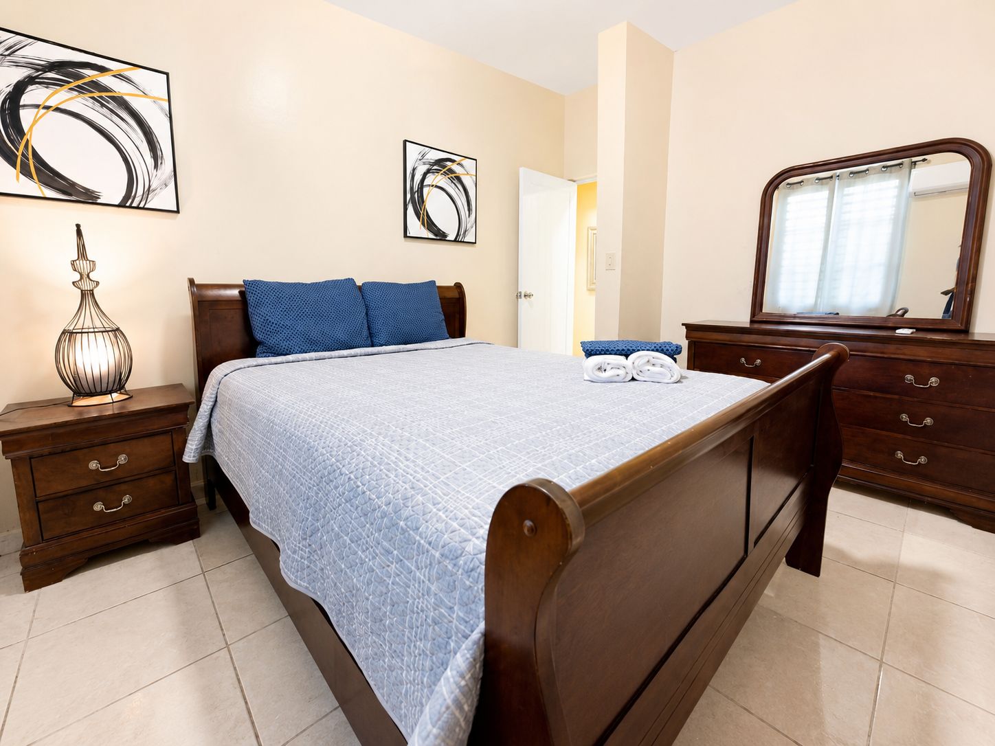 A bedroom with a bed, a nightstand, a dresser, a mirror, and two abstract paintings on the wall.
