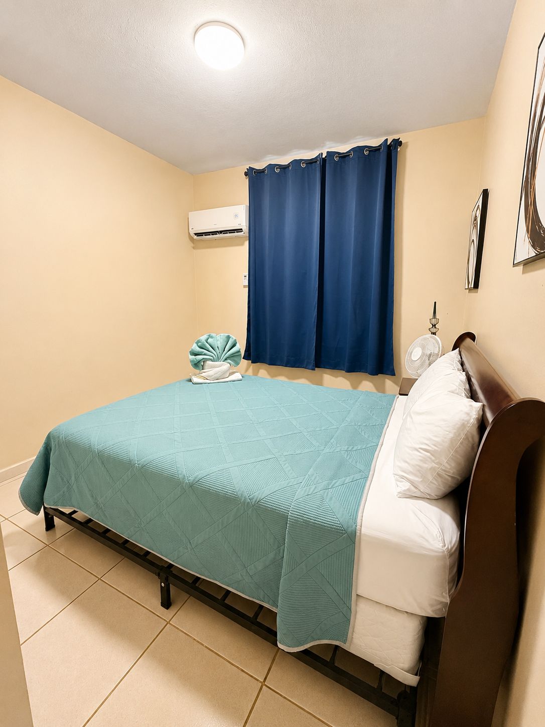Un dormitorio con una cama, aire acondicionado de pared, una ventana con cortinas y un ventilador de pie.