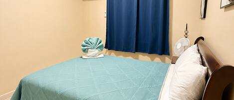 A bedroom with a bed, a wall-mounted air conditioner, a window with curtains, and a bedside fan.