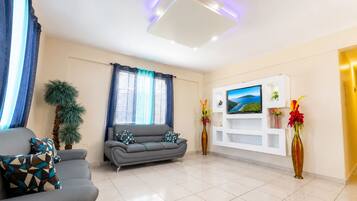 Un salón moderno con un sofá gris, un televisor de pantalla plana montado en una pared blanca, una planta en maceta y un jarrón con flores.