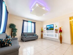 A modern living room with a grey sofa, a flat-screen TV mounted on a white wall, a potted plant, and a vase with flowers. - Colinas del Atlantico for 18 People (Isabela)