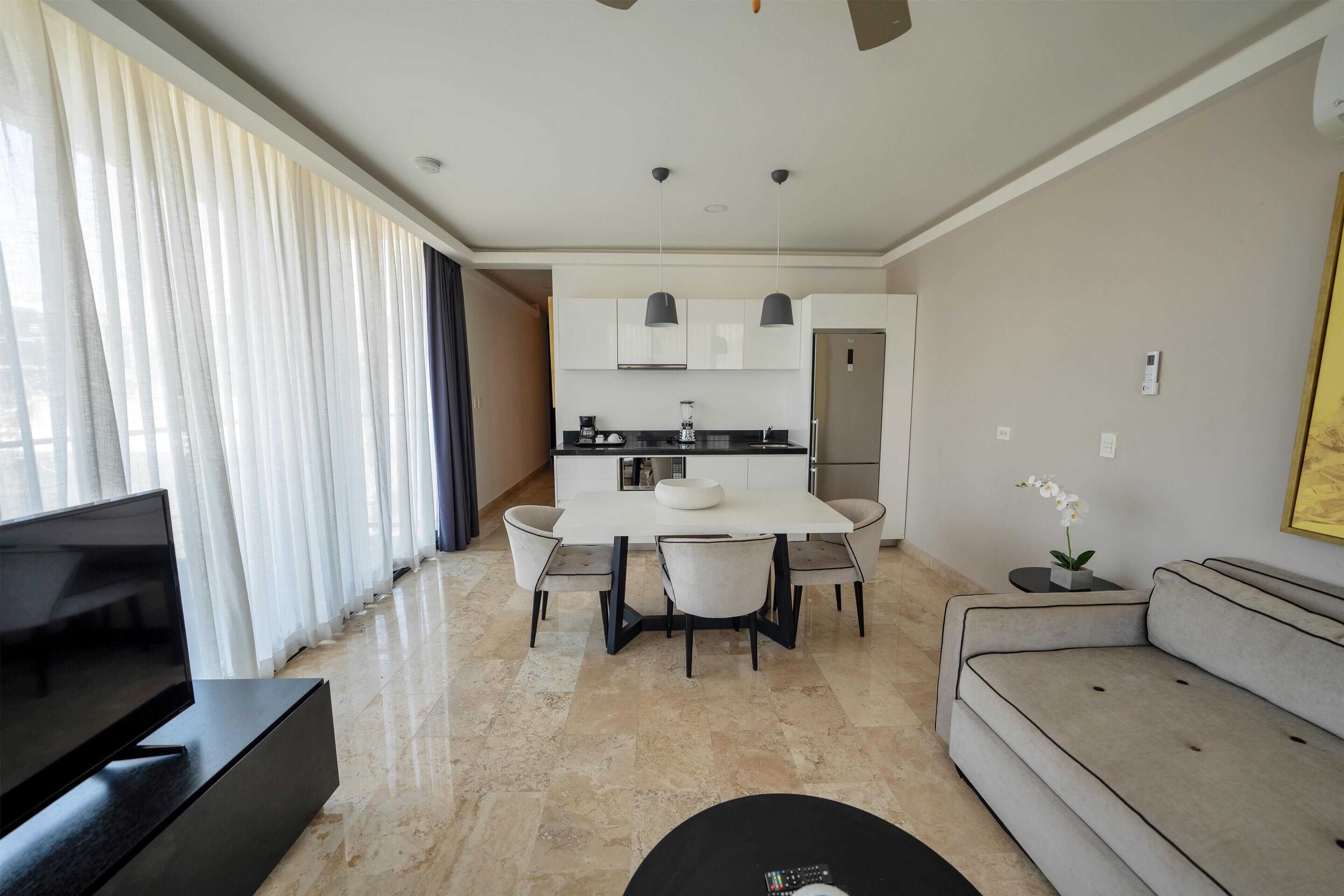 A modern living room with a flat-screen TV, a sofa, a dining table with chairs, and a kitchen area with cabinets and a refrigerator.