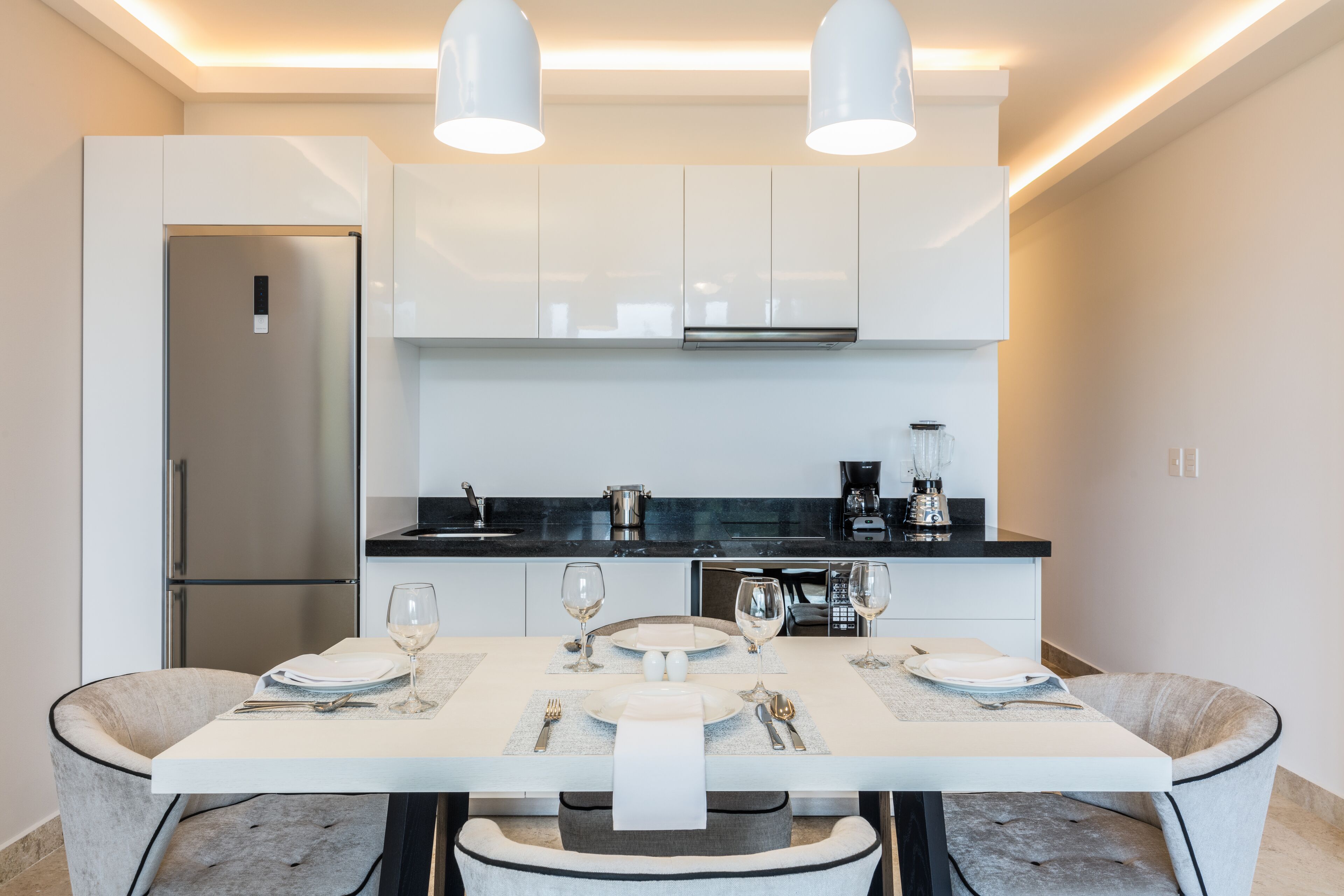 Une cuisine moderne avec des meubles blancs, un plan de travail noir et une table à manger dressée pour quatre personnes, avec des verres et des couverts.