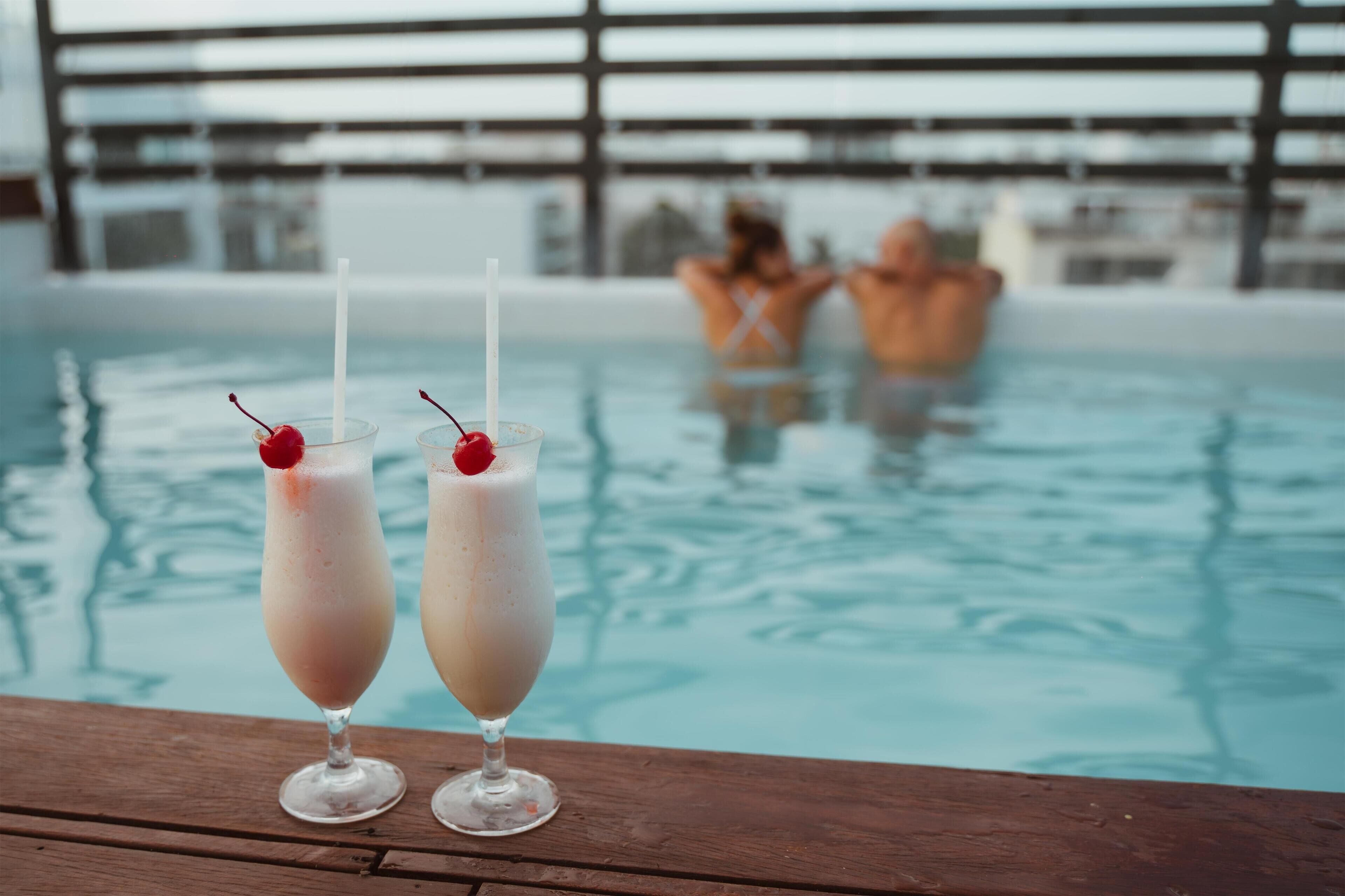 Dos bebidas con pajitas y cerezas sobre una superficie de madera junto a una piscina.