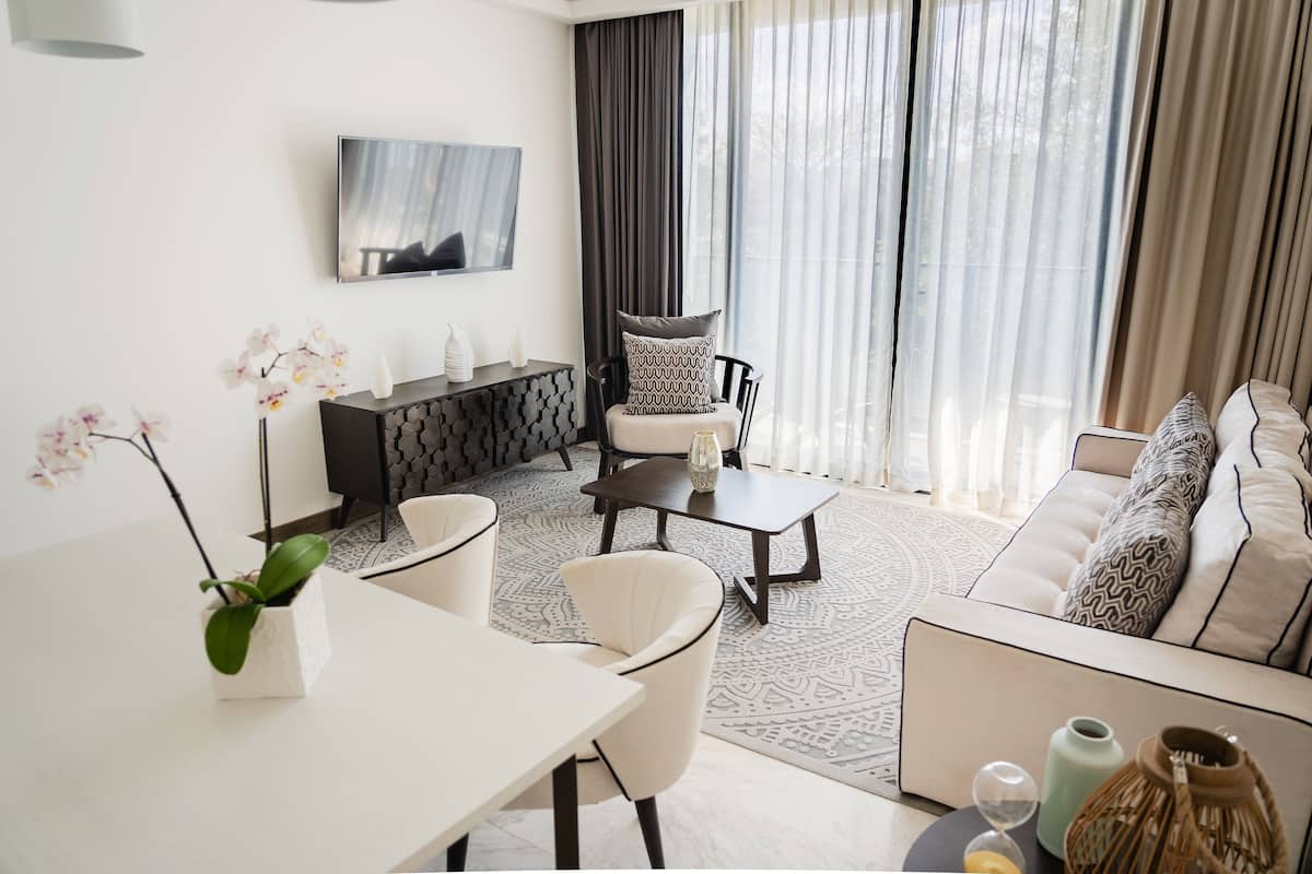 A modern living room with a sofa, armchairs, a coffee table, and a dining table. There is a TV mounted on the wall and a large window with curtains.