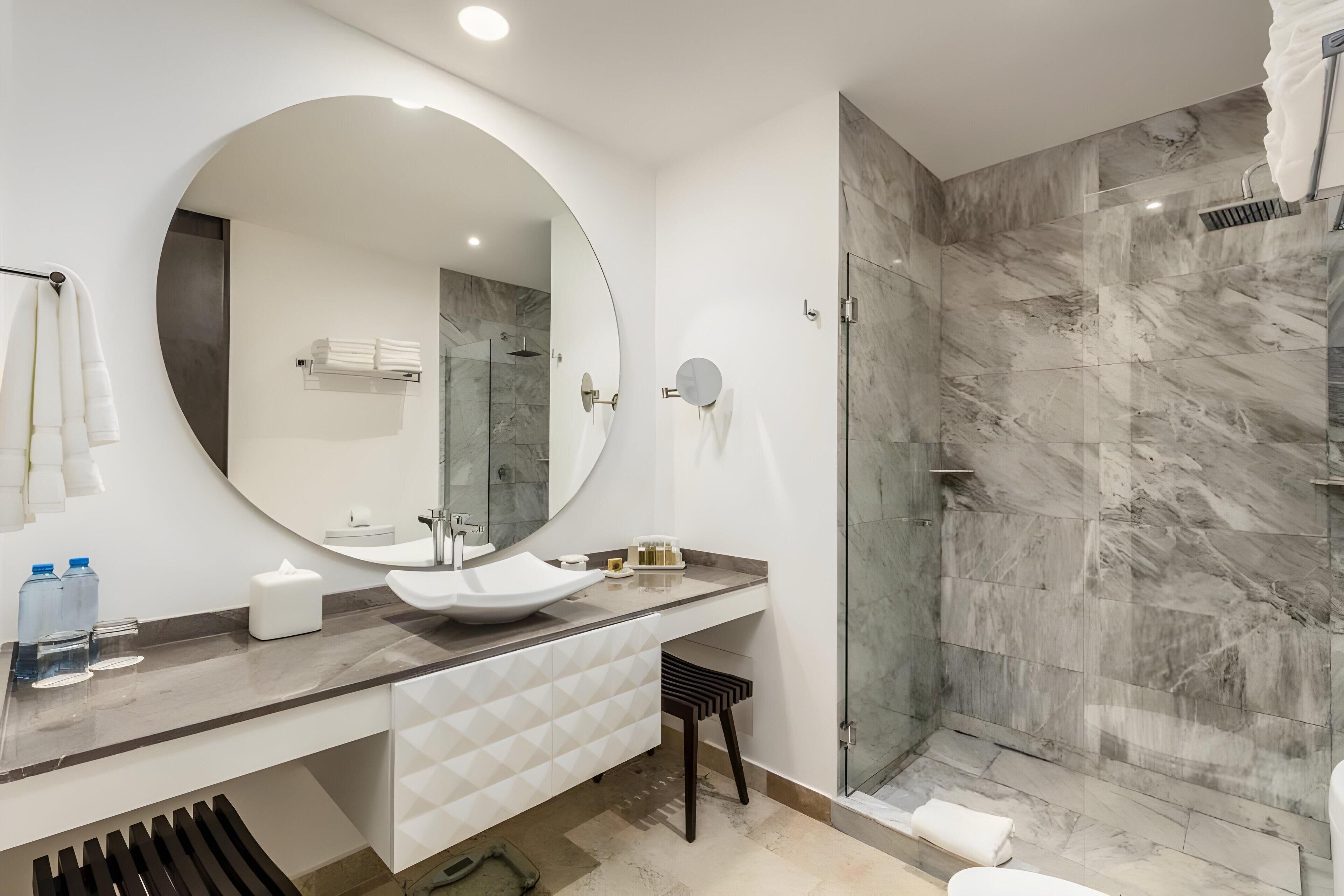Een moderne badkamer met een grote ronde spiegel, een wastafel op voet en een douche met glazen wanden.
