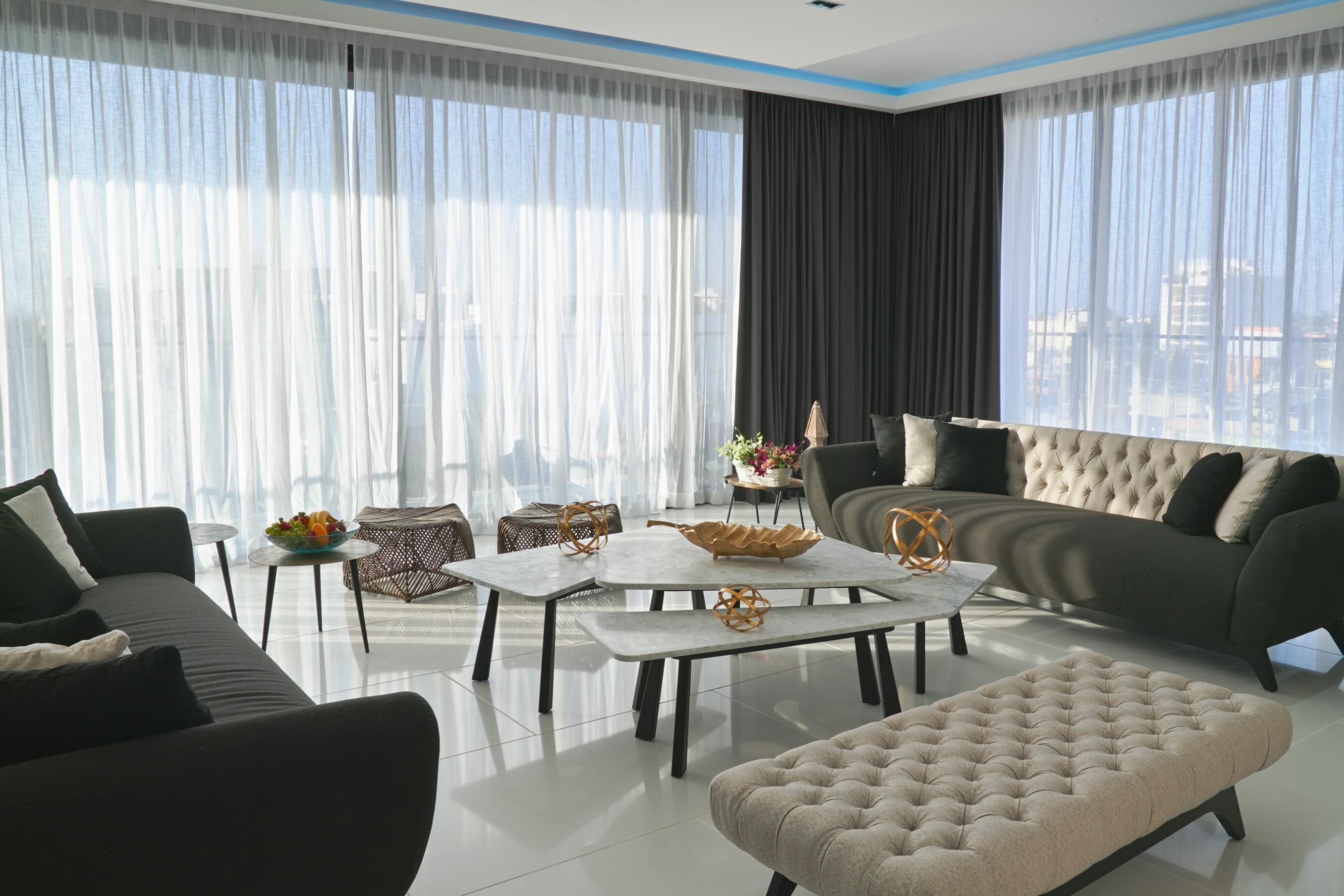 Una sala de estar moderna con sofá, reposapiés y mesa de centro, ventanas amplias con cortinas ligeras y vistas a la ciudad.