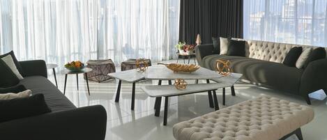 Una sala de estar moderna con sofá, reposapiés y mesa de centro, ventanas amplias con cortinas ligeras y vistas a la ciudad.