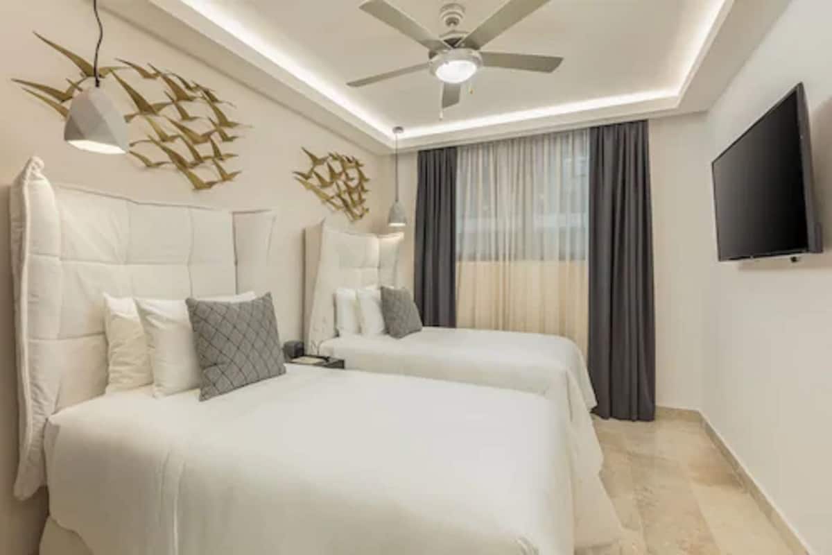 A modern hotel room with two beds, a ceiling fan, a flat-screen TV, and a decorative wall piece.