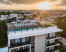 Un área de piscina en la azotea con sillones de descanso y sombrillas, rodeada de edificios modernos y vista al atardecer.