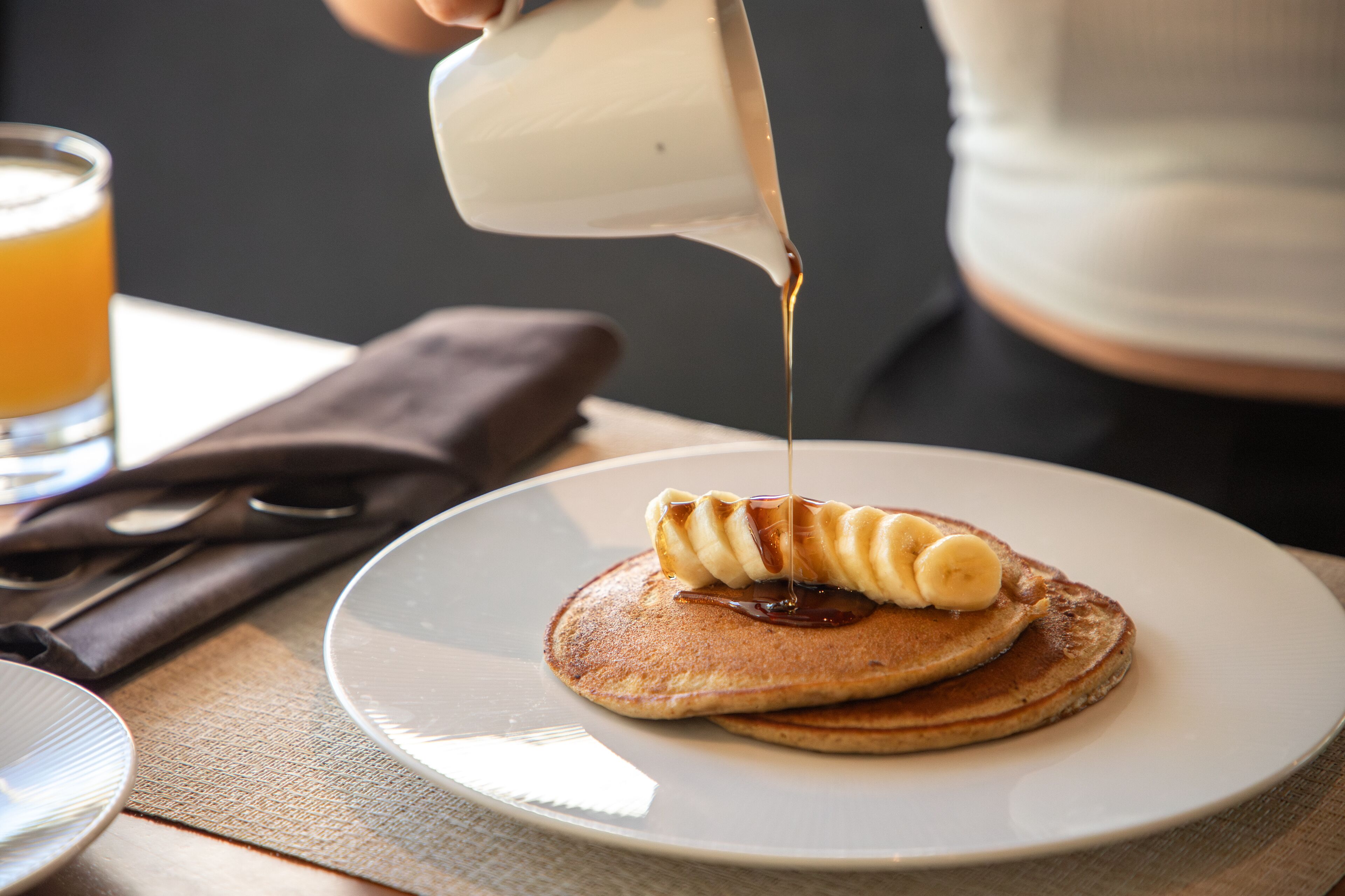Een bord met pannenkoeken, belegd met plakjes banaan en siroop die erover gegoten wordt.
