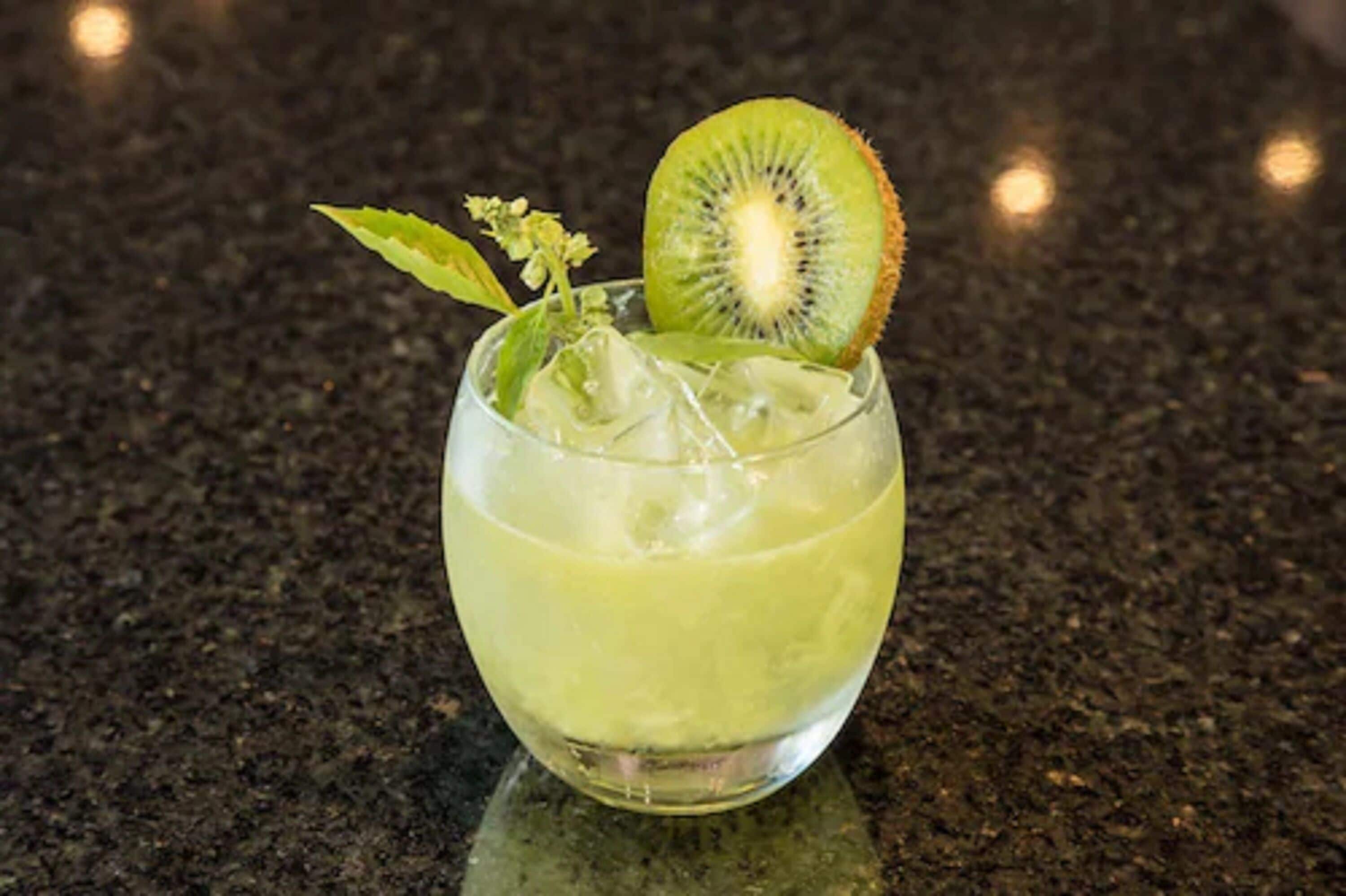 Un verre contenant une boisson verte, des glaçons, une tranche de kiwi et une feuille de menthe, posé sur un plan de travail foncé.
