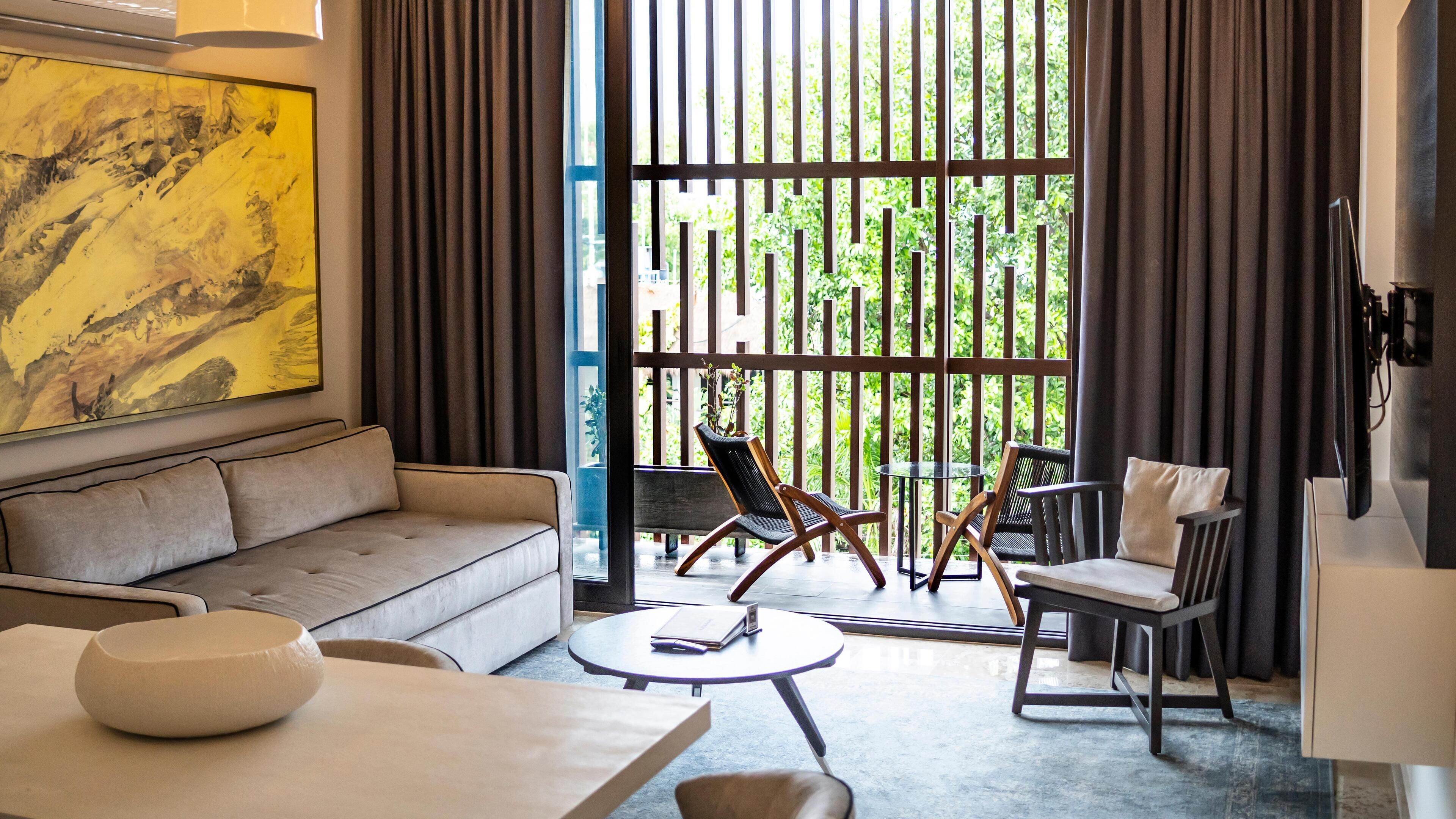A modern living room with a sofa, a round coffee table, and two chairs. There is a large window with vertical blinds, and a painting is visible on the wall.