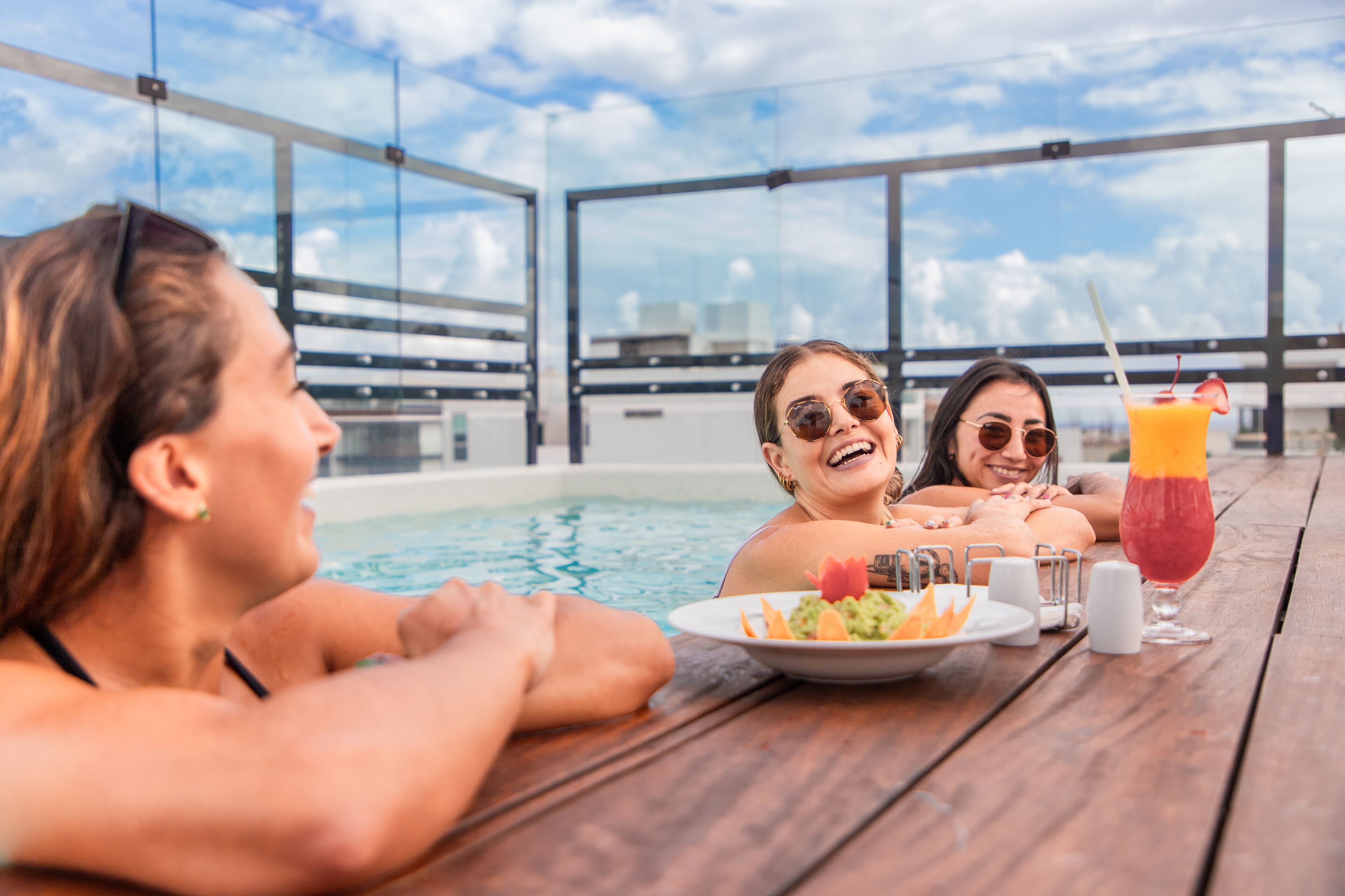 Une scène de bord de piscine avec une table en bois, un verre de jus d’orange et une assiette de nourriture.