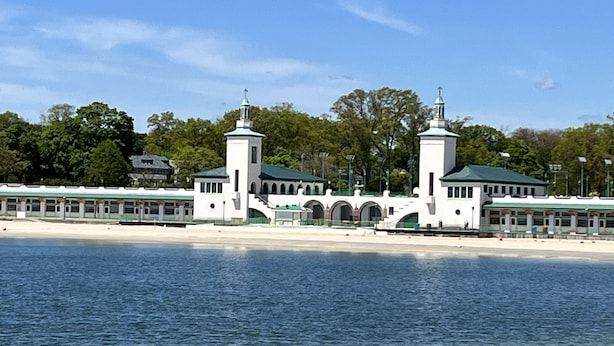 A white building with two towers and a green roof, located by a body of water.