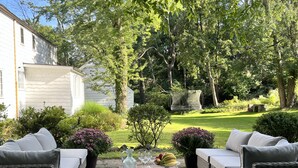 An outdoor patio with wicker furniture, a table with a pitcher and fruit, and a garden with various plants and a swing chair.