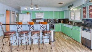 Une cuisine avec des meubles de couleur verte, un réfrigérateur blanc, un four à micro-ondes et un évier. On y trouve quatre tabourets de bar en bois, avec des assises marron.