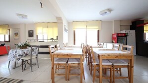 Uma área de refeições com mesas e cadeiras de madeira, uma cozinha com geladeira e uma máquina de Coca-Cola vermelha.