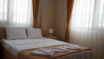 A neatly made bed with white linens and a brown throw blanket, a bedside table with a lamp, and sheer curtains.