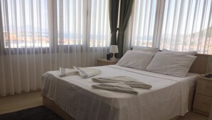 Cama bem arrumada com lençóis brancos, mesa de cabeceira com abajur e vista para o exterior através de cortinas leves.