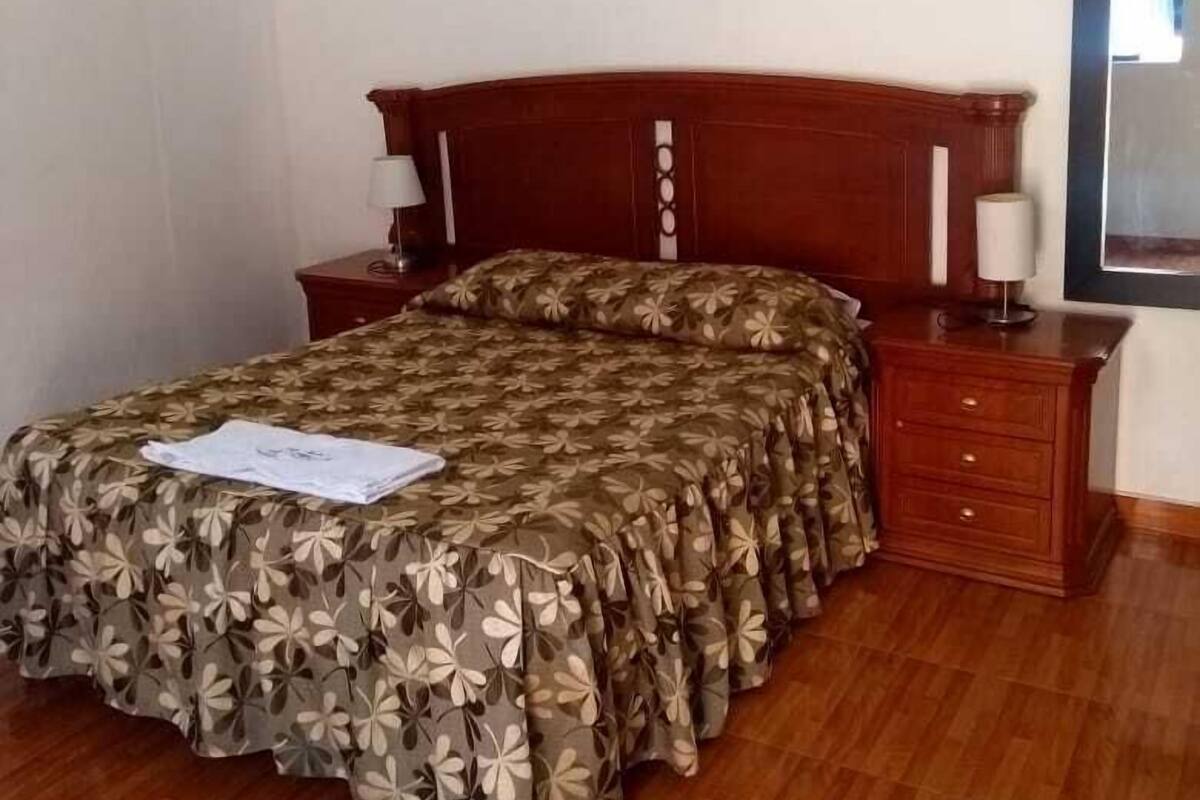 Ein Hotelzimmer mit einem hölzernen Bett, einem blumigen Überwurf, zwei Nachttischen mit Lampen sowie einem Bild an der Wand.