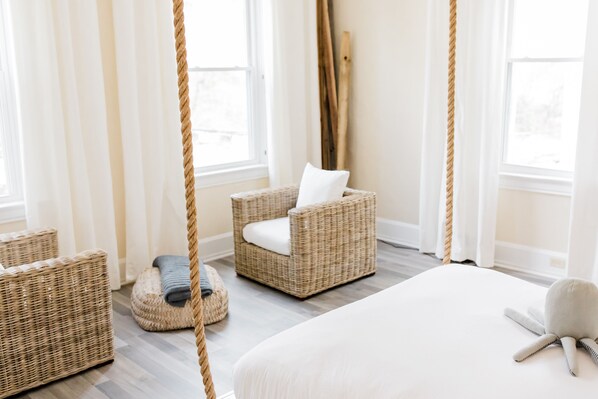 Une pièce avec un grand lit, un fauteuil en osier avec un coussin blanc, un petit pouf rond avec une couverture grise et une balançoire en corde.
