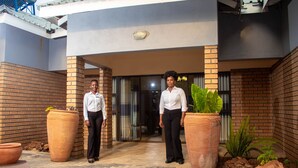 Two individuals standing in front of a building with brick walls and large potted plants.